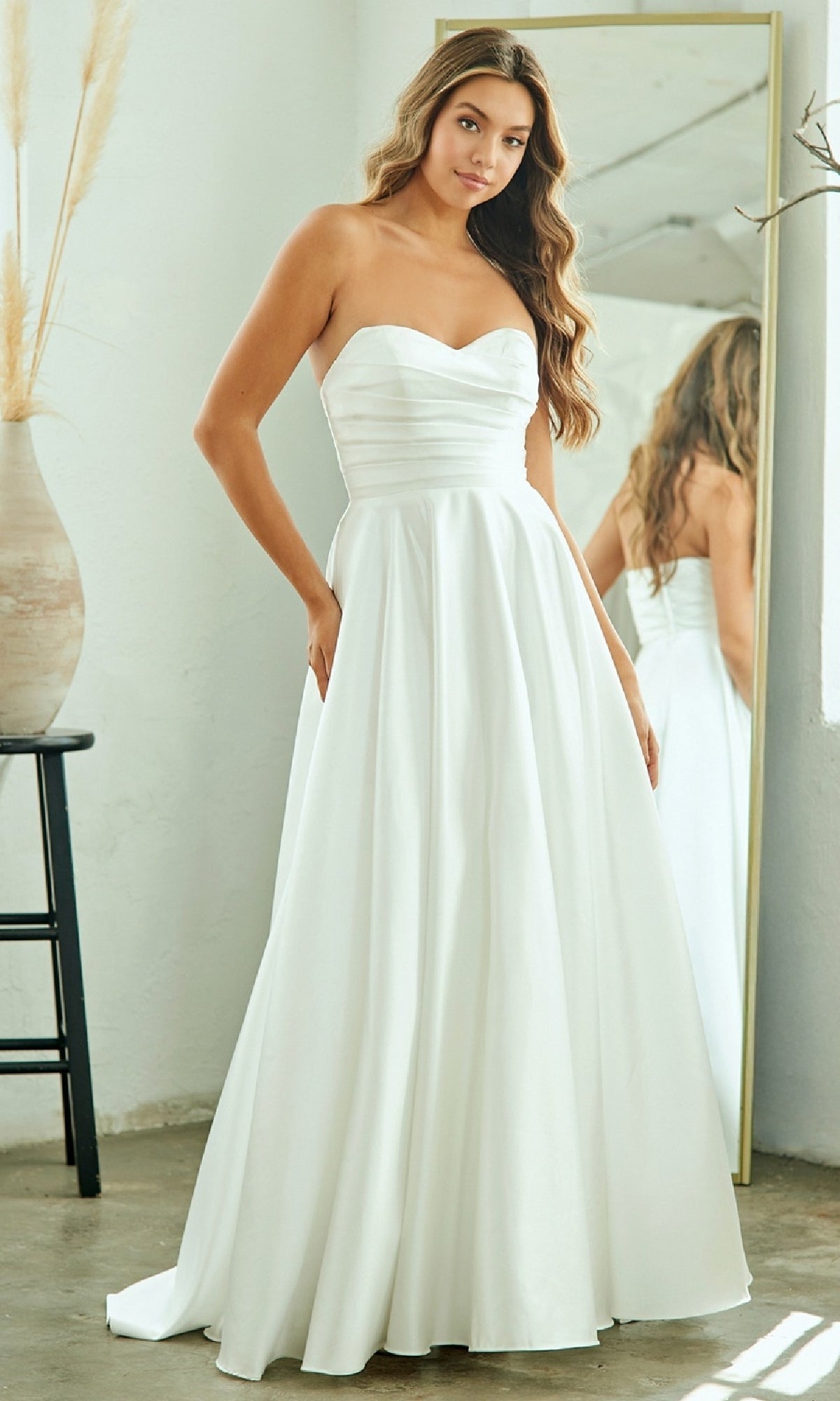 A woman stands in front of a mirror in a minimalist room wearing the Amelia Couture 5076 Classic Simple White Prom Dress, featuring a strapless fitted bodice and a full skirt.